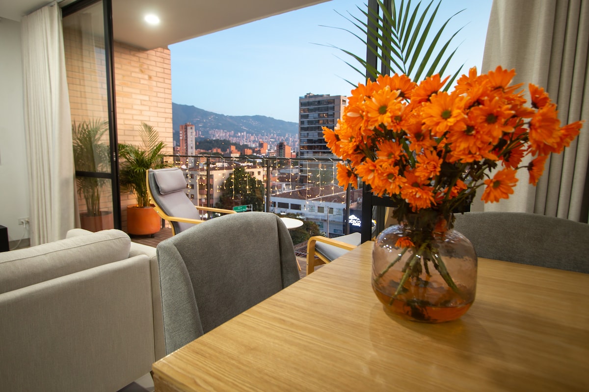 A light-filled living space features a wooden dining table alongside modern chairs. A vase of vibrant orange flowers adds a splash of color. Large glass doors lead to a balcony, showcasing urban views and distant mountains under a twilight sky.