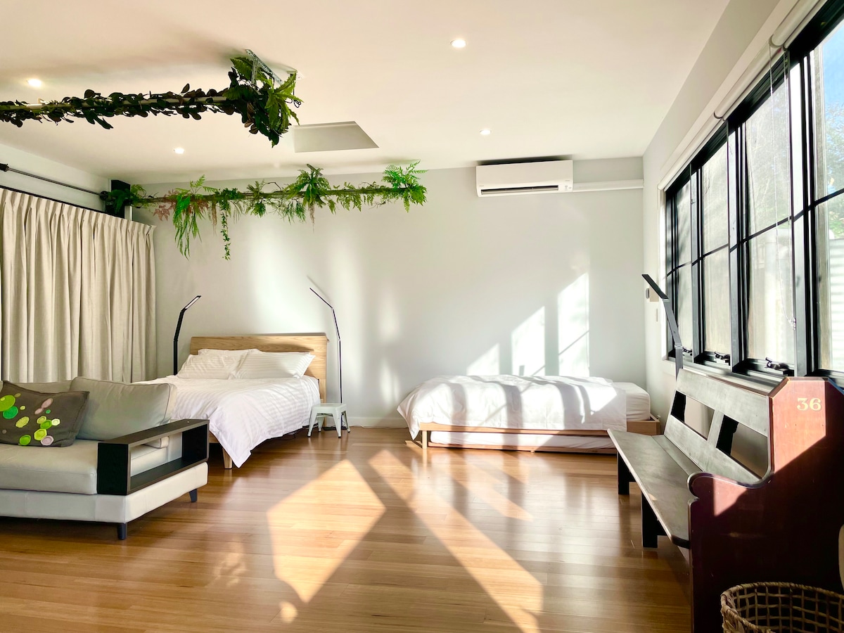 A spacious studio features a queen bed and a single trundle bed, arranged near large windows that allow natural light to fill the room. A wooden bench is positioned against one wall, and decorative plants hang from the ceiling, enhancing the airy environment.