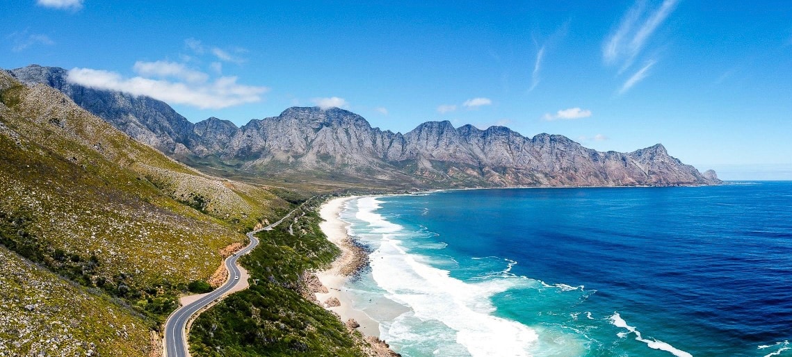 A panoramic view showcases a winding coastal road alongside a serene ocean. Rugged mountains rise majestically in the background, while gentle waves are seen lapping against the shore. The sky is bright blue, with a few wispy clouds scattered above.