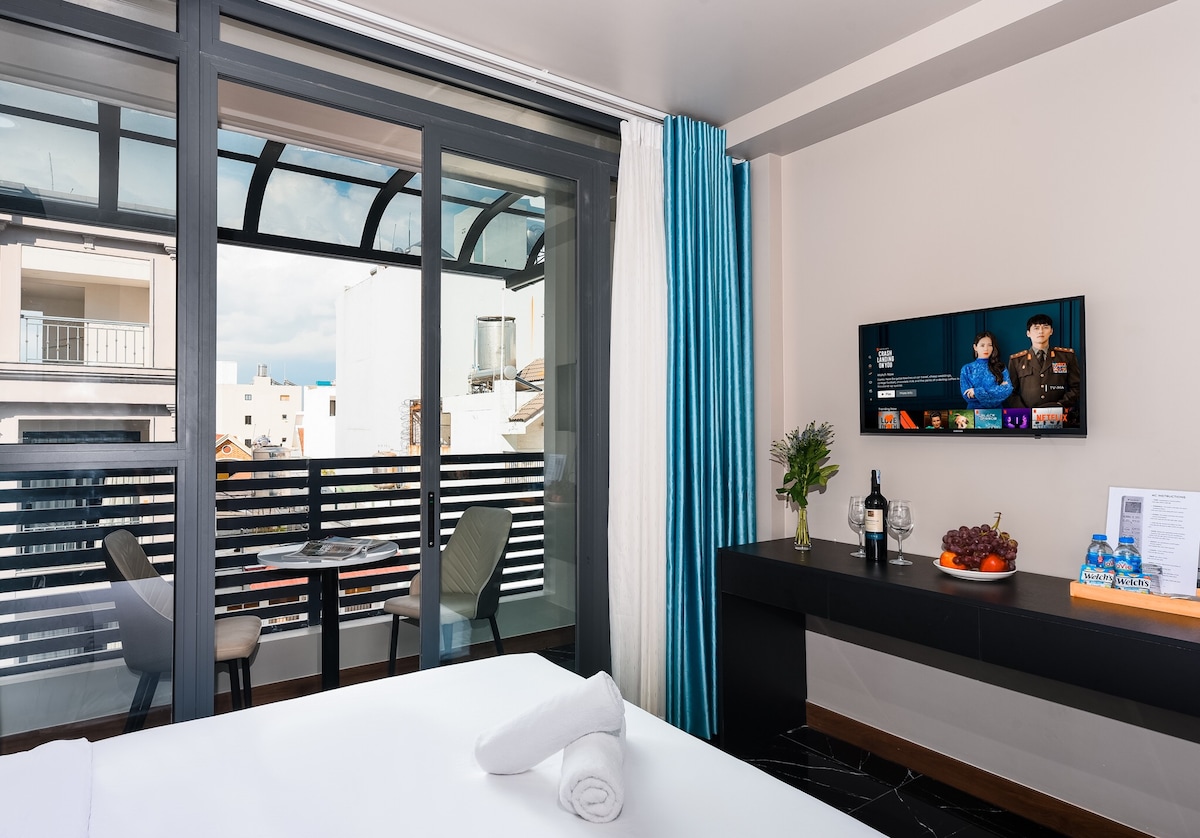 A modern interior is observed with a spacious bed in the foreground and a stylish table set on a balcony. A smart TV displays entertainment options. Large glass doors allow natural light to fill the room and provide a view of the outdoors.