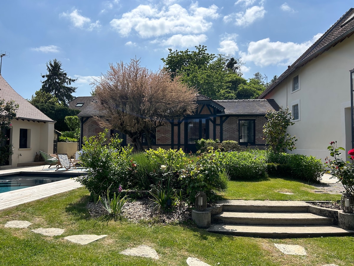 The image presents a peaceful garden area featuring a mix of greenery and blooming flowers. A stone pathway leads toward a charming cottage, with a wooden deck in front of a pool. A few lounge chairs are set beside the pool, under a clear blue sky.