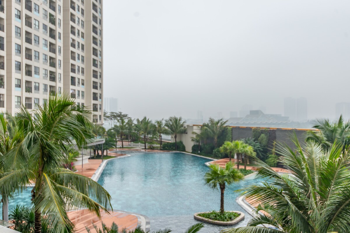 The image depicts a tranquil outdoor pool area surrounded by palm trees. The pool features a smooth surface reflecting the surrounding greenery and buildings. A misty backdrop adds a serene ambiance to the scene, creating a peaceful atmosphere for relaxation.