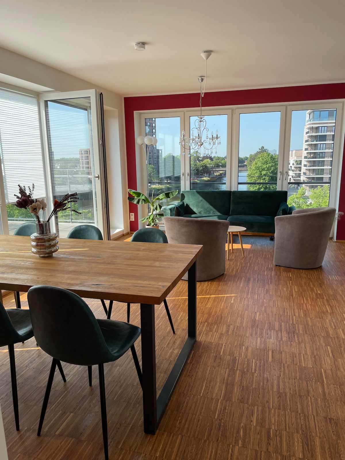 A bright living space features large glass doors opening to a view of the river and surrounding greenery. A wooden dining table is surrounded by four dark chairs. Two gray armchairs and a green sofa create a comfortable seating area, complemented by a decorative chandelier overhead.