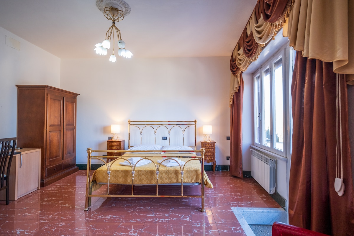 The room features a spacious gold-colored bed framed in brass, complemented by two bedside lamps. A wooden wardrobe and a small table are present, along with a chair. Natural light enters through multiple windows adorned with curtains, showcasing the tranquil atmosphere.