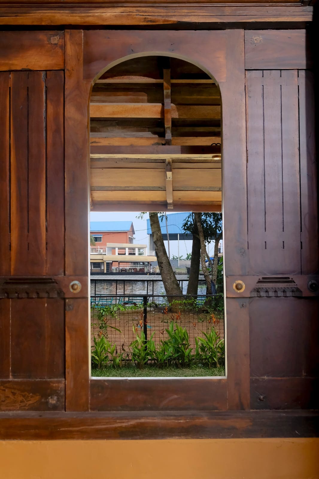 A wooden cabinet with an arched mirror reflects a view of a green outdoor space. Visible through the mirror are trees and a fenced area, providing a sense of the surrounding environment, while the cabinet’s design adds a touch of rustic character.