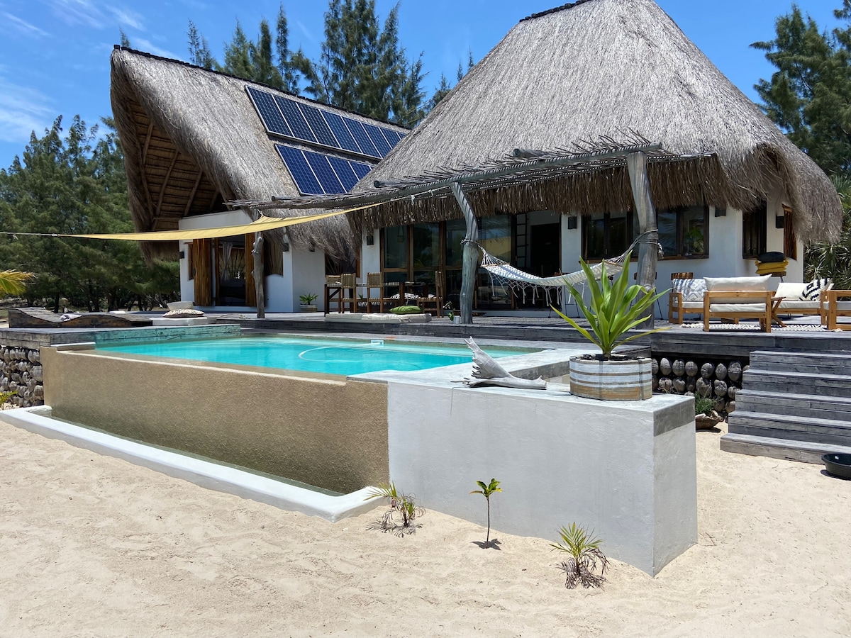 A spacious infinity pool is set against a sandy backdrop, bordered by a white wall and adorned with potted plants. The adjoining thatched-roof villa features solar panels and large windows that invite natural light. Sun loungers and outdoor seating areas enhance the relaxing atmosphere.