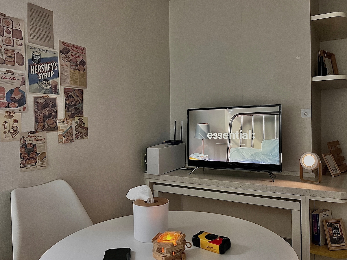 A compact living area features a round table with a soft light source and a television displaying a still image. Walls are decorated with various recipe cards and posters, contributing to a cozy atmosphere. A tissue box and a small decorative item are placed on the table.