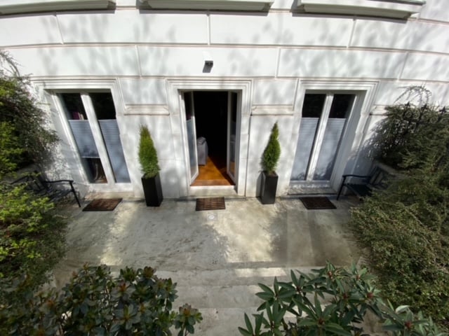 The entrance of the apartment is flanked by two planters with leafy greenery. A pair of black chairs is positioned on the patio, while the double doors lead into the interior, allowing natural light to fill the space.