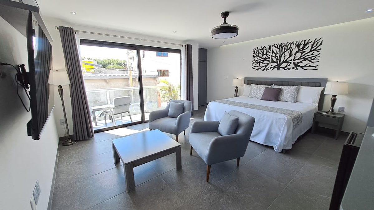 A spacious bedroom showcases a large bed with decorative pillows, flanked by two bedside lamps. Two gray chairs and a small table are positioned by a sliding glass door, which opens to a balcony. Natural light enhances the modern decor and neutral tones.