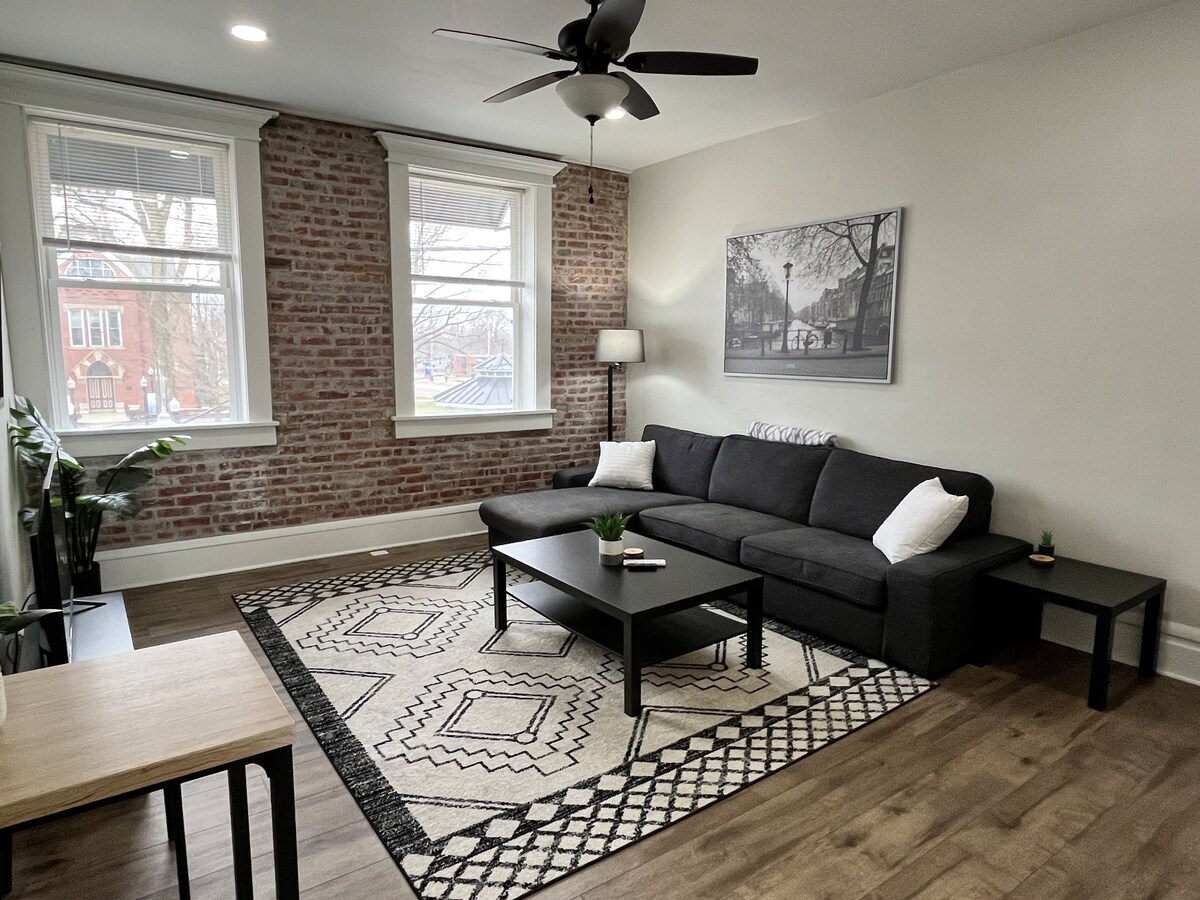 A spacious living area is presented with a dark sectional sofa and a rectangular coffee table. Natural light enters through two large windows, illuminating exposed brick walls. A patterned area rug anchors the seating, complemented by indoor plants and a side table.