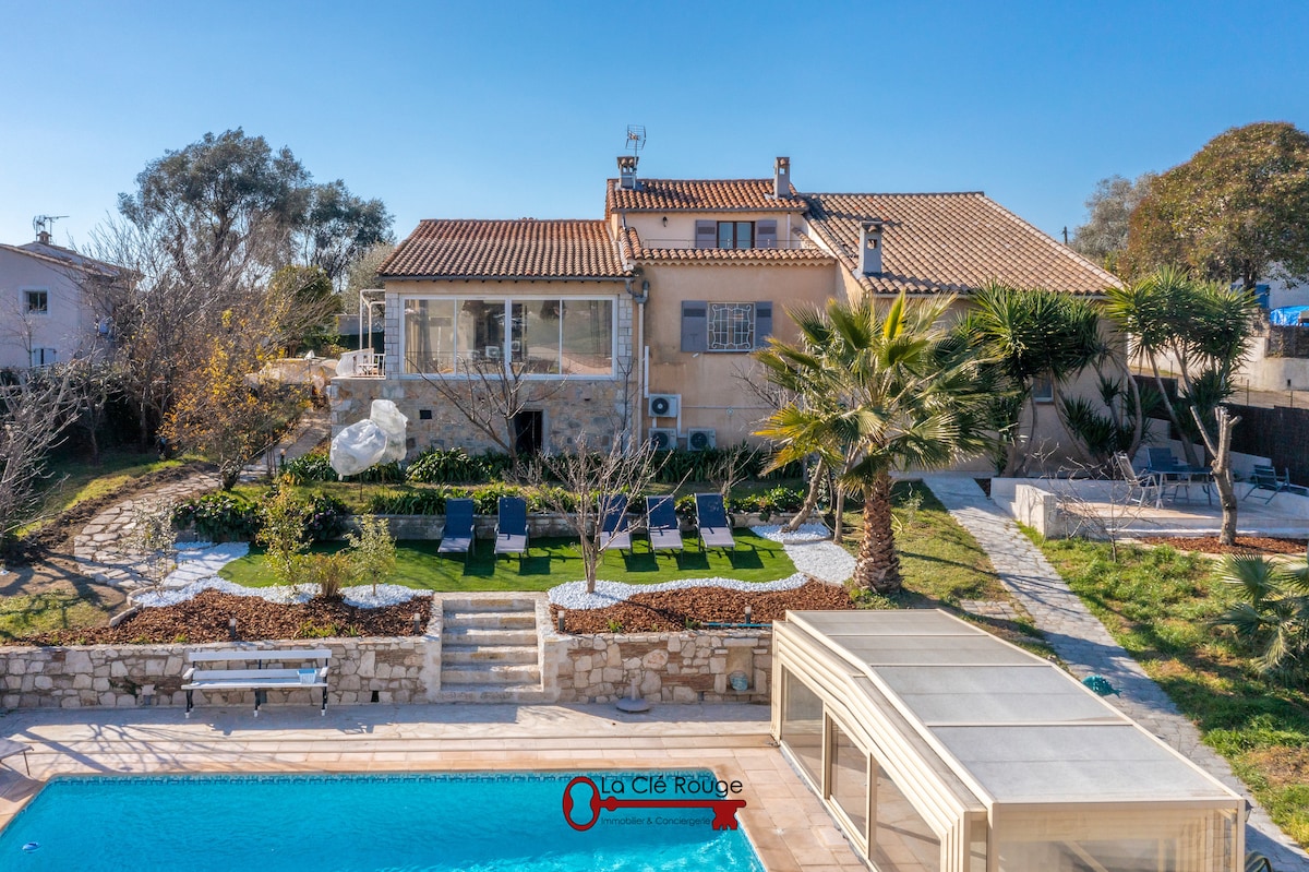 A spacious villa is set amidst landscaped gardens, featuring a tranquil pool in the foreground. Sun loungers are neatly arranged around the pool, while palm trees and stone pathways frame the home's exterior. The stone facade and terracotta roof complement the bright blue sky.
