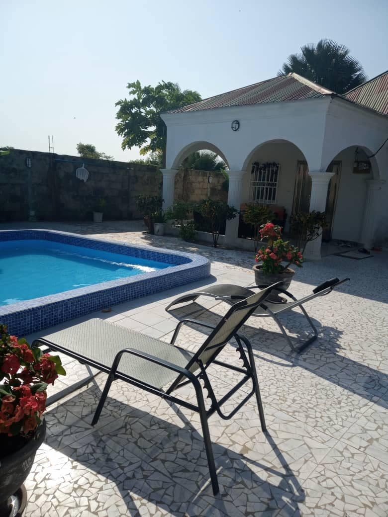 A serene outdoor space includes a sparkling pool surrounded by tiled flooring. Two lounge chairs are positioned beside vibrant potted plants, creating a relaxing environment. A white, arched structure is visible in the background, contributing to the overall tranquil setting.