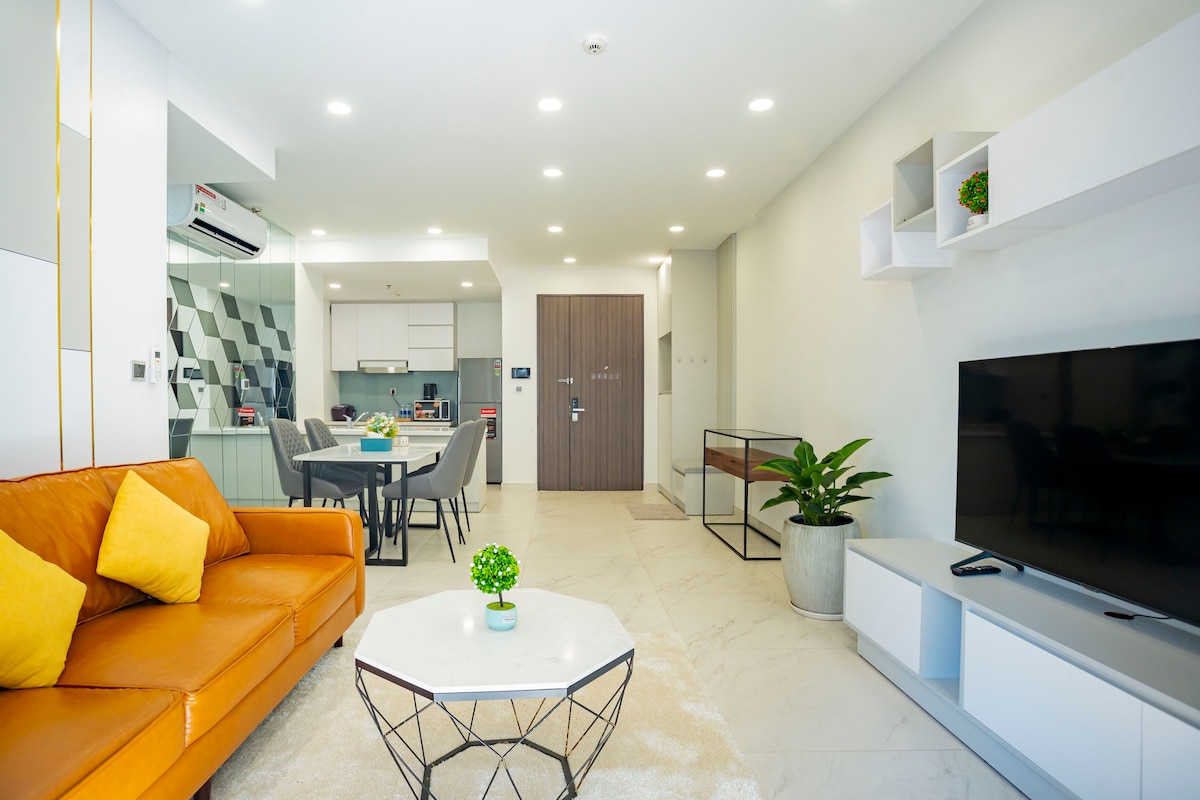 A modern living area is showcased with a large orange sofa, complemented by decorative pillows. A round coffee table sits in front, and a green plant brings a touch of nature. The kitchen and dining area are visible in the background, with sleek appliances and light fixtures.