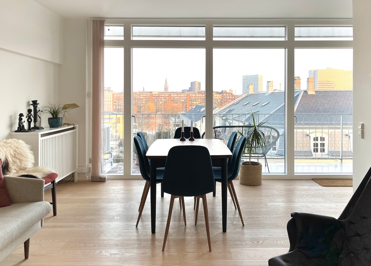 A dining area features a wooden table surrounded by black chairs. Large windows provide a view of the city skyline, with warm lights visible in the distance. Soft furnishings, including a cozy throw and decorative items, complement the space.