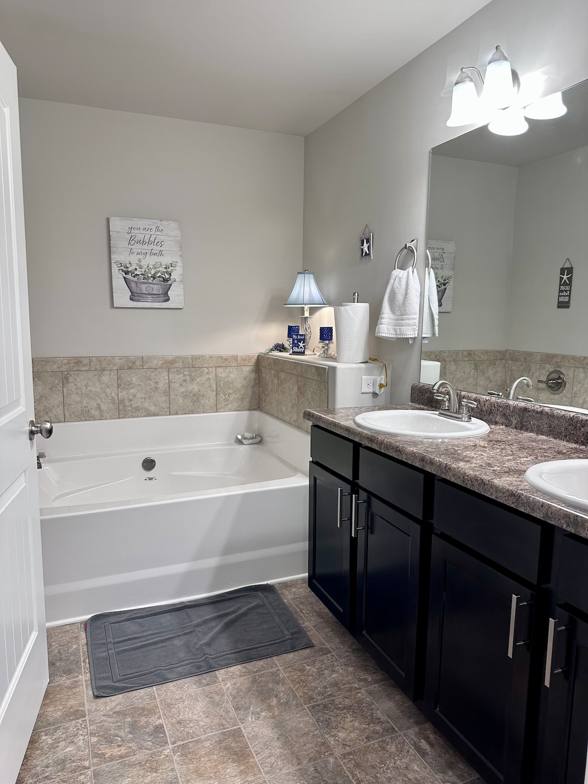 A well-appointed bathroom features dual sinks with sleek dark cabinetry and a large mirror. A light blue lamp provides soft illumination, complemented by a white towel rack. A spacious soaking tub is positioned against a tiled wall, with neutral-colored flooring enhancing the clean aesthetic.