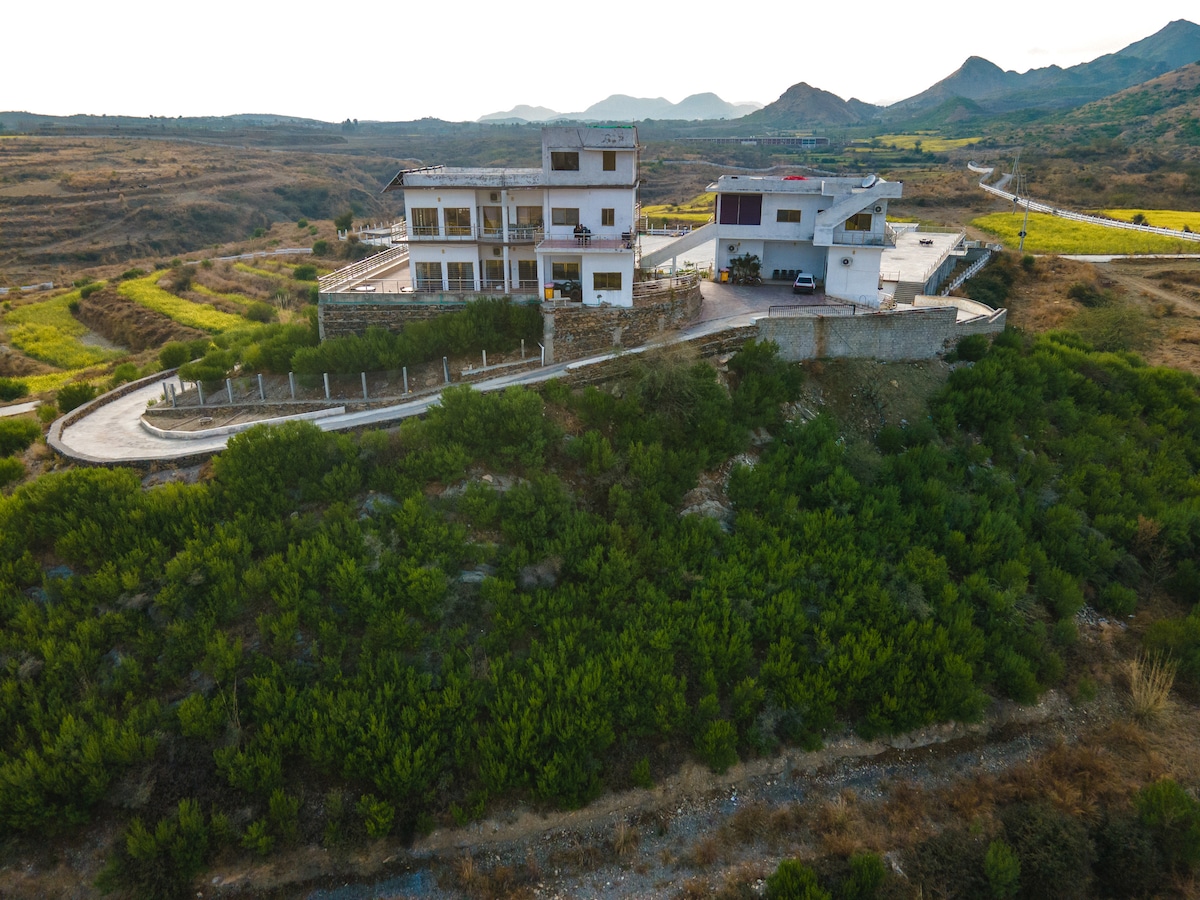 Aerial view of the resort, featuring two distinct buildings surrounded by lush greenery and rolling hills. Scenic landscapes stretch into the distance, highlighting the natural beauty of the area. The road leading to the resort is visible, creating a sense of accessibility.