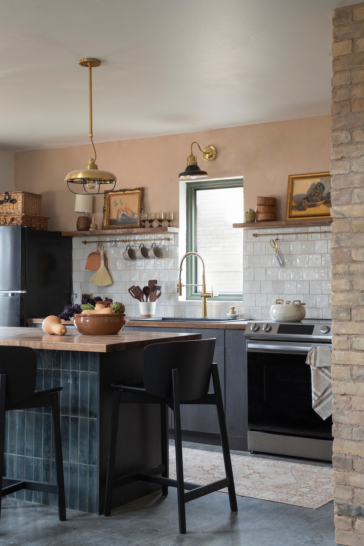 The kitchen features modern appliances, including a stove and refrigerator. A wooden countertop is complemented by seating at two high stools. Wall-mounted shelves hold various kitchen items, while decorative artwork enhances the overall design. Natural light enters through a window framed by neutral walls.