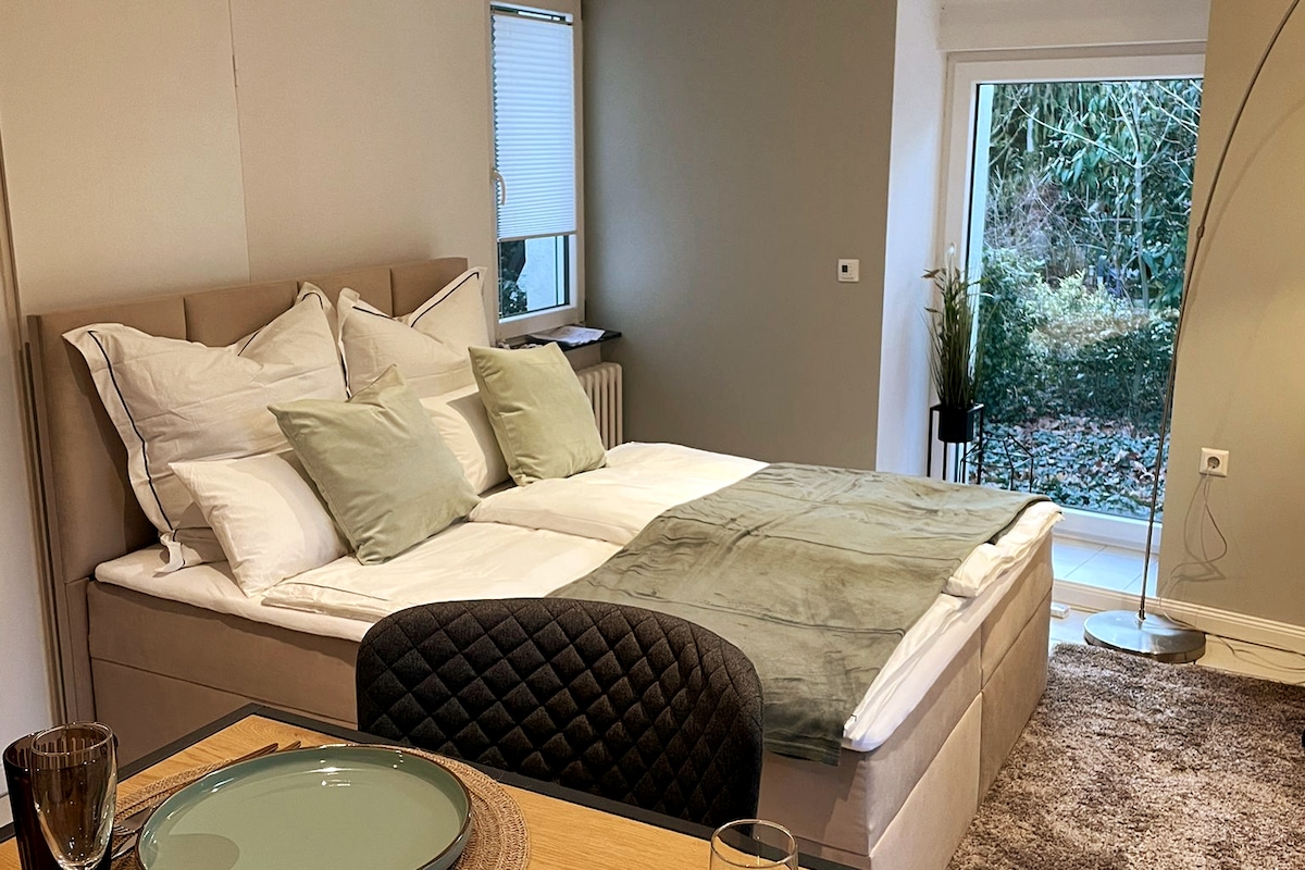 A spacious bed is centered in the room, adorned with soft gray and white linens and supportive pillows. Natural light enters through a nearby window, illuminating a view of the greenery outside. A dining area is positioned in the foreground, featuring a dark, quilted chair and elegant table settings.