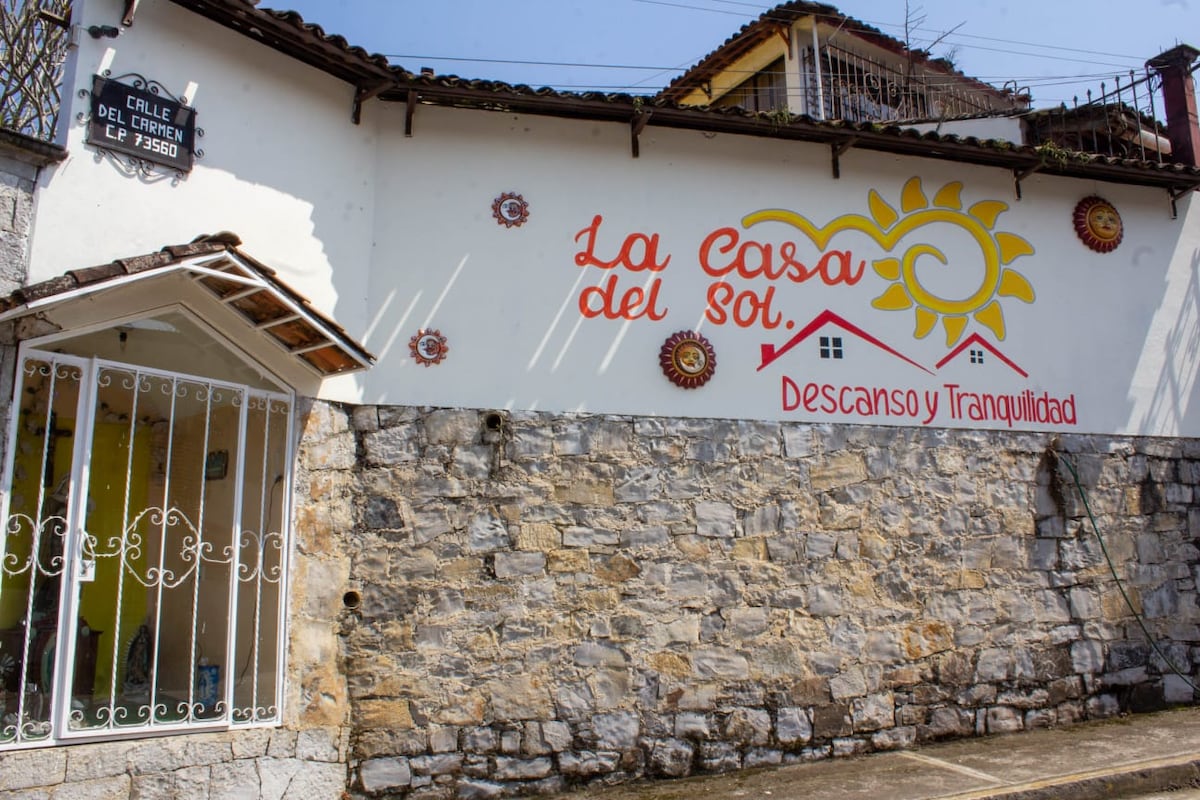 A stone wall is adorned with a vibrant mural featuring a sun and the text 'La Casa del Sol. Descanso y Tranquilidad.' A gated entrance with a simple roof is visible, contributing to a welcoming facade.
