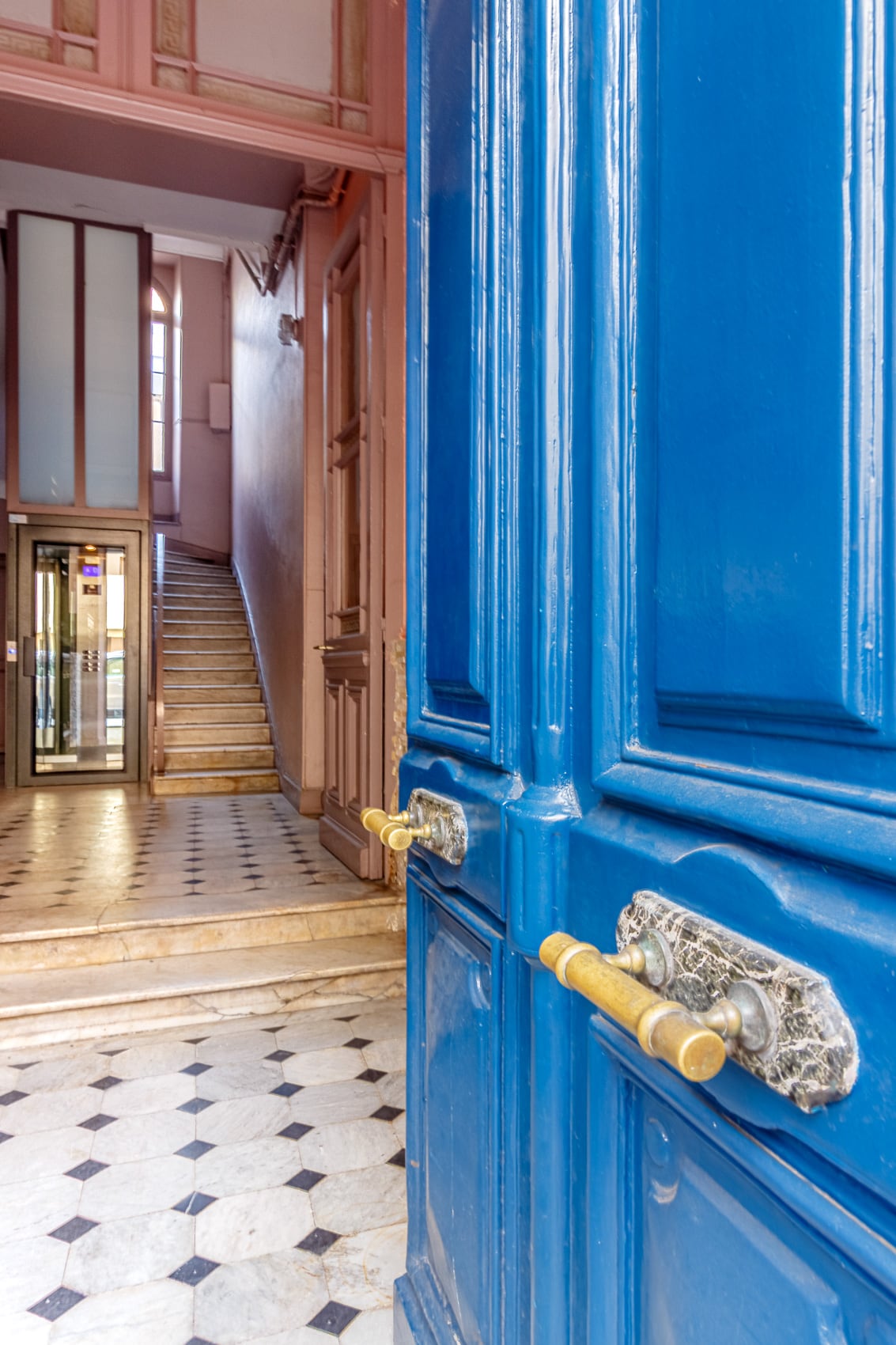 A vibrant blue entrance door opens to reveal a marble hallway. An elevator is visible at the far end, adjacent to a staircase with elegant steps. The walls display subtle pastel colors, enhancing the welcoming ambiance of the entrance.
