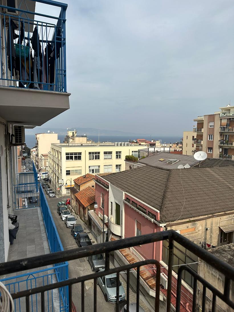 A balcony is shown overlooking the neighborhood, revealing a view of streets lined with buildings and parked cars. In the distance, the sea is faintly visible beneath a cloudy sky. The balcony features blue railings, adding a pop of color to the scene.