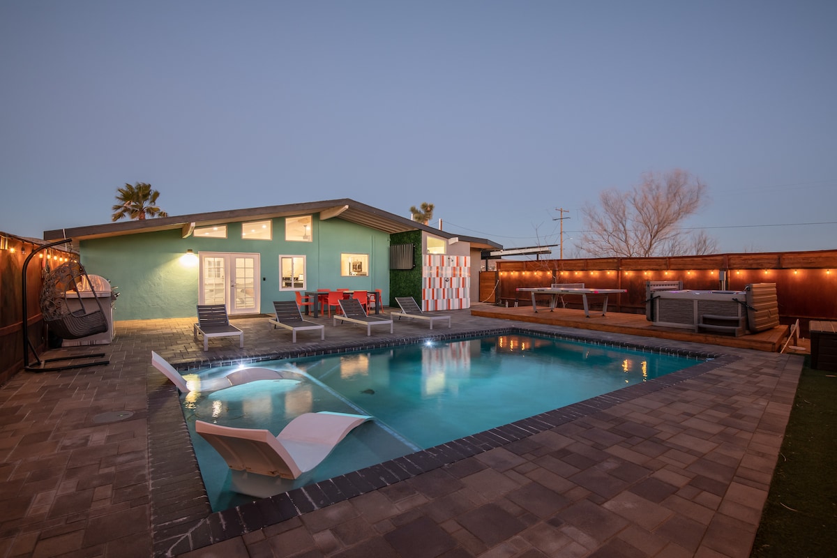 An inviting backyard features a clear swimming pool surrounded by a spacious patio. Sun loungers are arranged near the water's edge, and a shaded seating area offers additional comfort. Soft outdoor lighting sets a relaxing ambiance as day transitions to night.