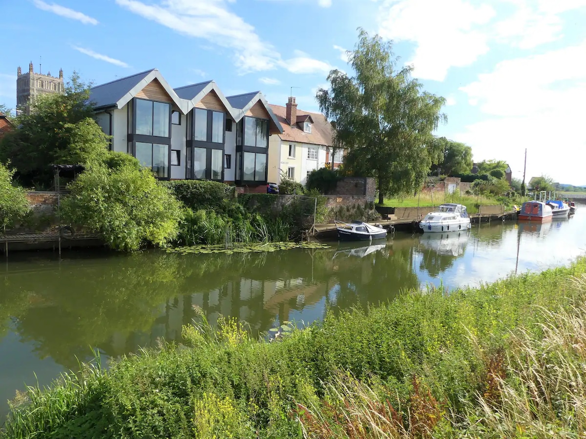 The image captures a scenic view of the River Avon with a variety of boats moored along the bank. On the opposite side, a charming two-story Victorian home is visible alongside modern buildings, framed by lush greenery and distant trees under a clear blue sky.