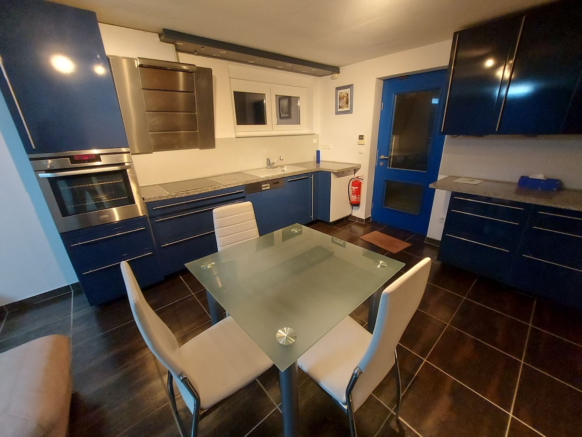 A modern kitchen is displayed, featuring deep blue cabinetry and a sleek glass-top dining table surrounded by four white chairs. Stainless steel appliances are integrated into the design, while a window lets in natural light. The floor is finished with dark tiles for a contemporary look.
