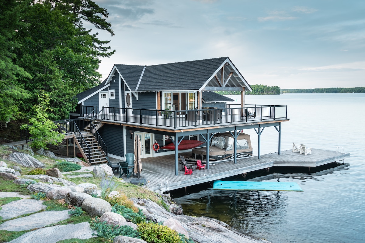 The boat house features a spacious deck overlooking calm waters, with ample seating and a covered boat slip. Natural stone steps lead to the water's edge, framed by lush greenery and rock formations, creating a serene and serene waterfront environment.