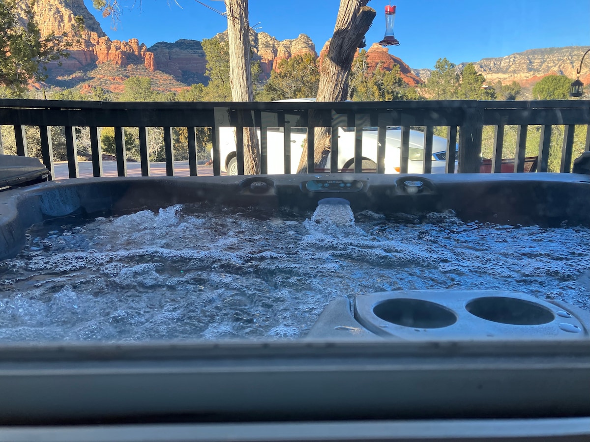 A hot tub, with bubbling water, is positioned on a deck overlooking stunning red rock formations. A glimpse of a vehicle and trees is visible in the background. A clear blue sky enhances the serene outdoor setting.