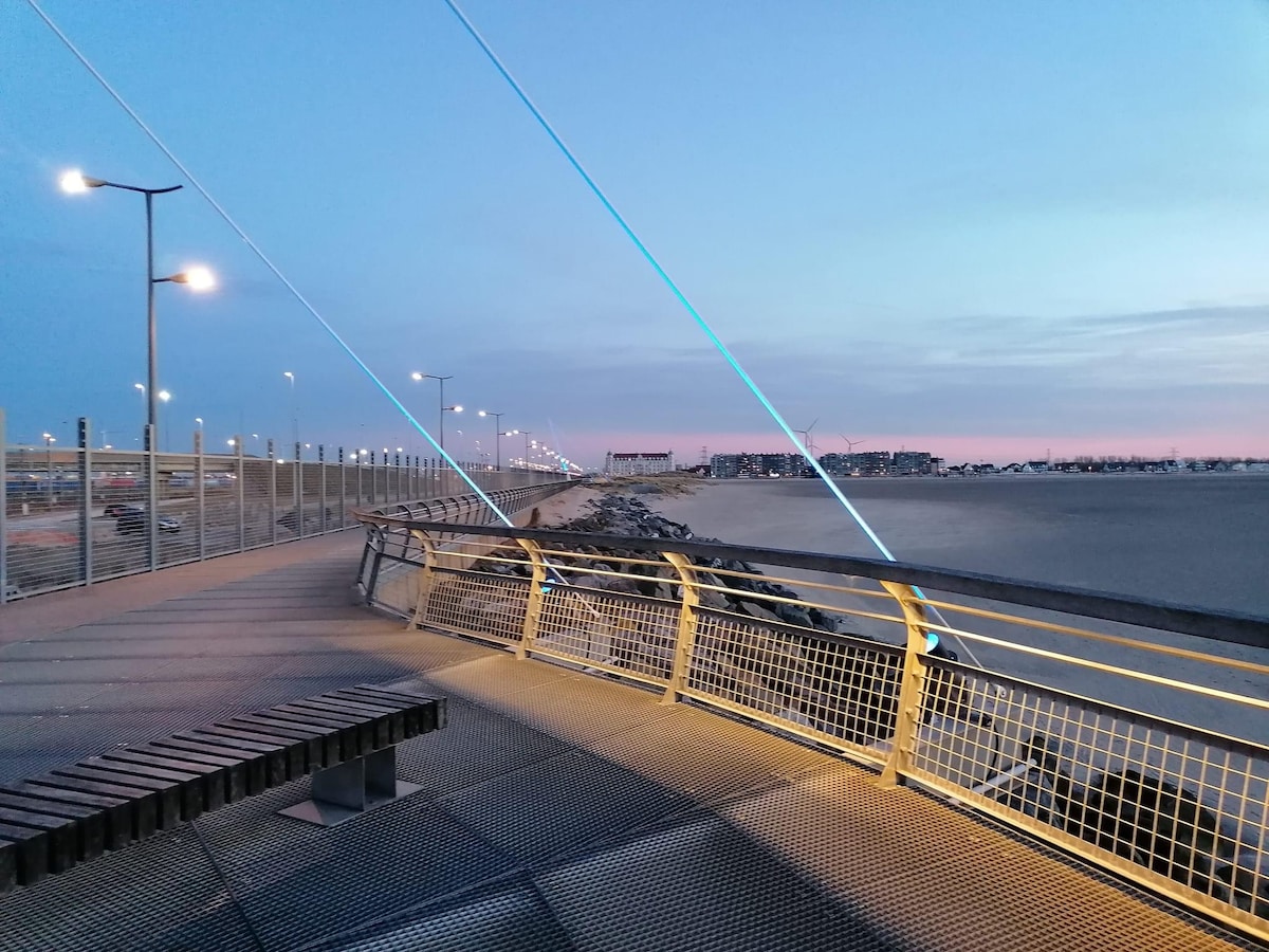 An illuminated promenade is presented along the sandy beach at dusk, featuring a modern railing and benches. Soft lighting enhances the surroundings, while distant buildings and wind turbines are visible under a colorful sky transitioning from blue to pink.