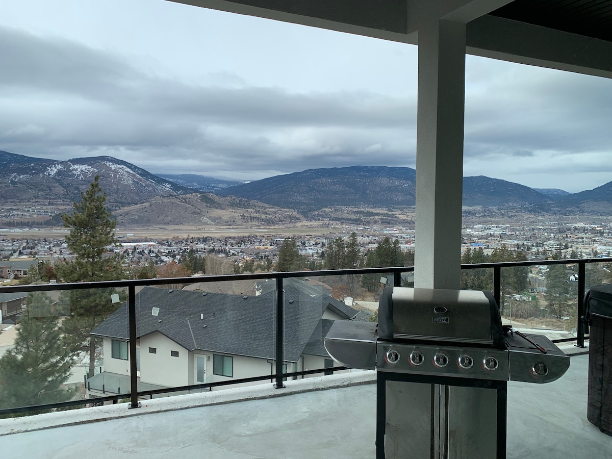 An outdoor patio is visible, featuring a barbecue grill positioned near the railing. The expansive view includes rolling hills and a distant town, under a cloudy sky. The modern architecture integrates well with the surrounding landscape.