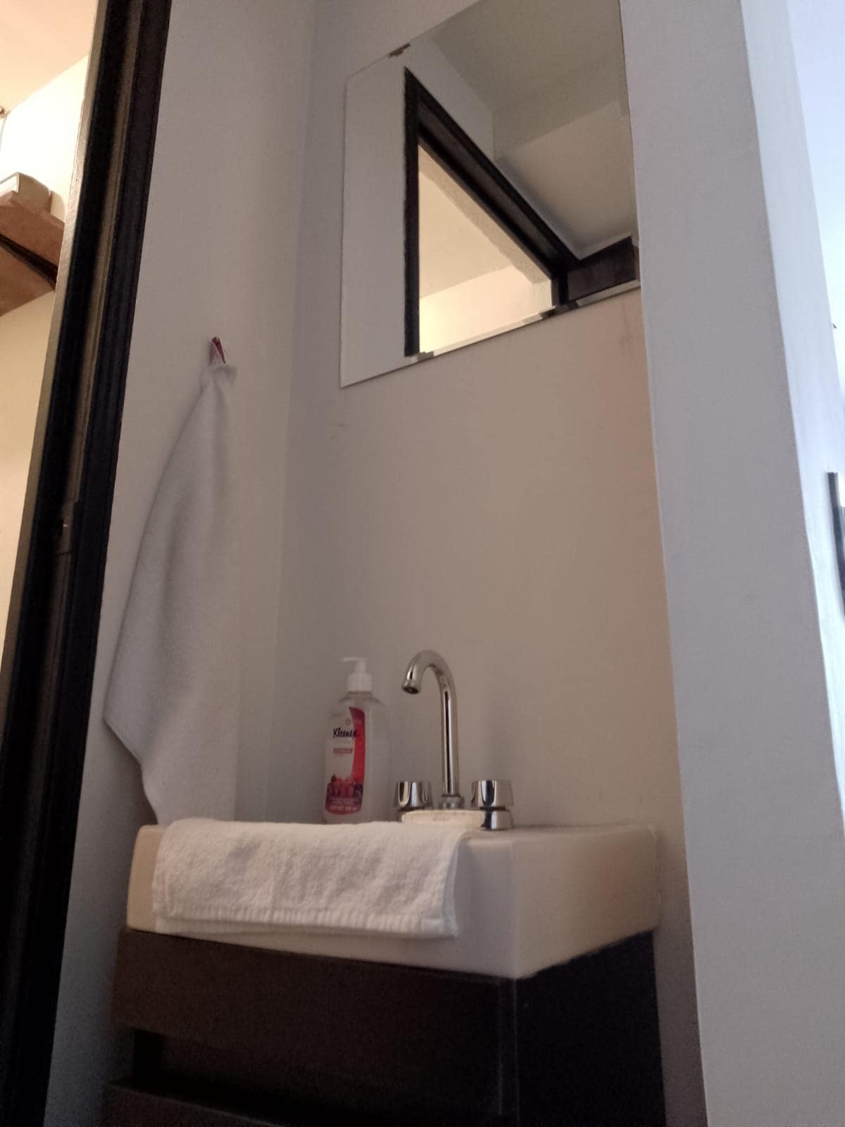 A compact bathroom area features a minimalist sink with a modern faucet. A white towel is neatly arranged beside a liquid soap dispenser on the dark countertop. A mirror is positioned above the sink, reflecting the surrounding light.