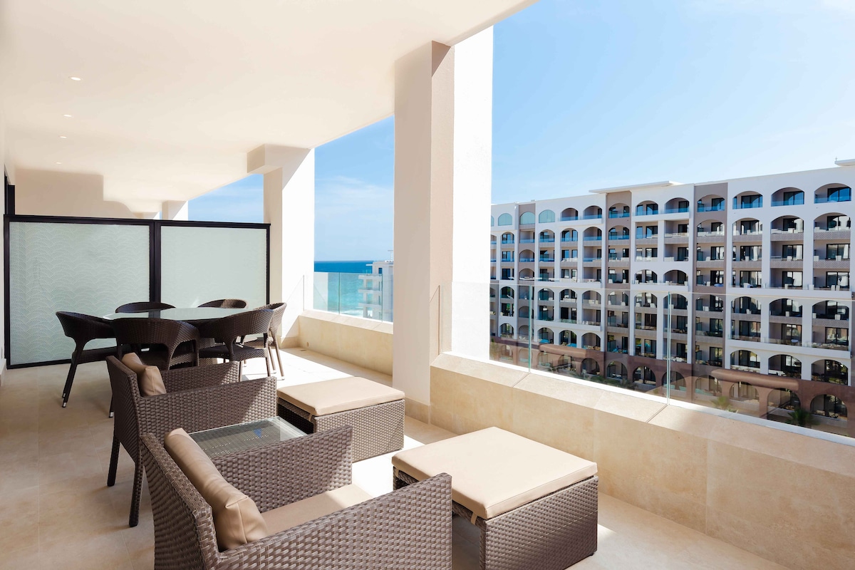 An outdoor terrace is furnished with dark wicker seating and beige cushions, offering a comfortable space to relax. A dining table with chairs complements the area, with the ocean visible in the background, framed by large glass panels and a clear blue sky.