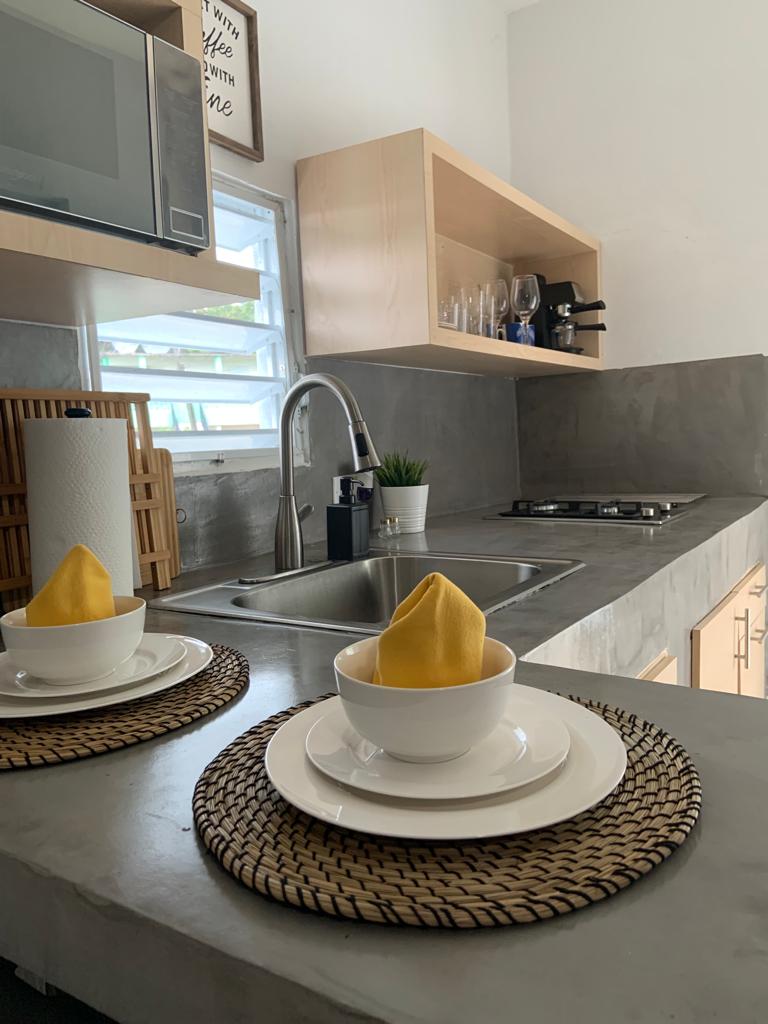 The kitchen features a modern design with concrete countertops and light wood cabinetry. A double sink is positioned beneath a window, with kitchen essentials neatly arranged nearby. Two bowls are presented on woven placemats, adding a touch of warmth to the space.