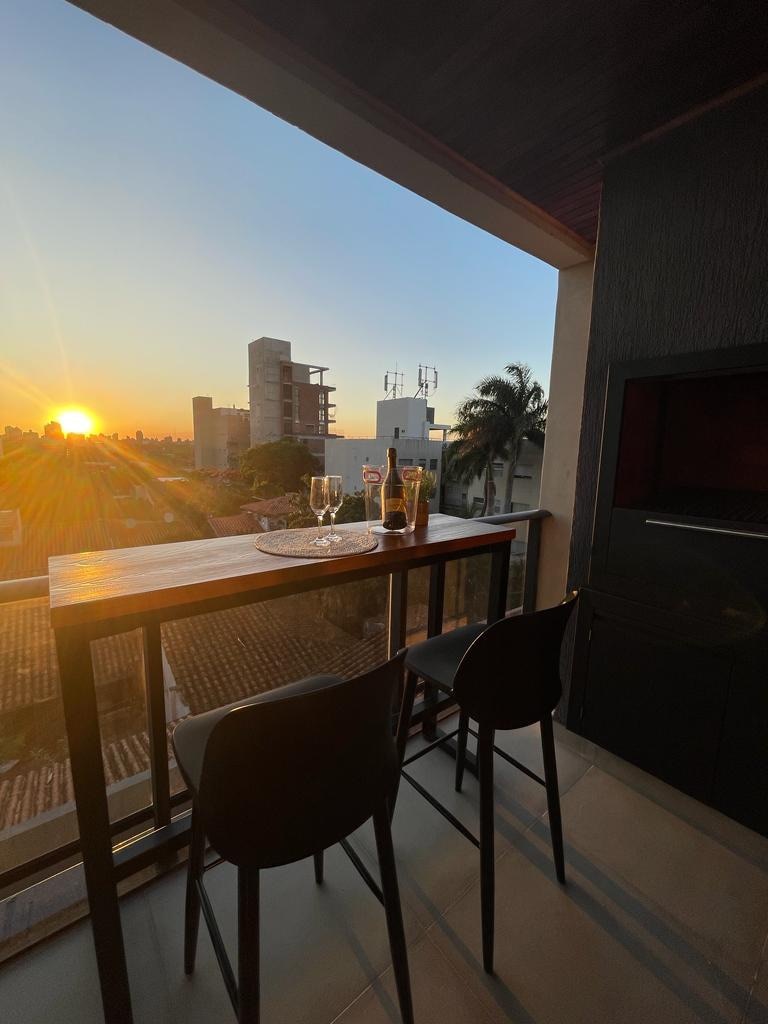A balcony offers a bar-height table set for two, featuring sparkling glasses and a bottle. The warm glow of sunset creates an inviting backdrop over a cityscape, while palm trees gently sway in the distance.