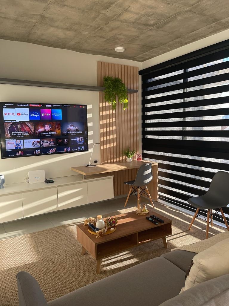 A modern living area is highlighted by a wall-mounted television displaying various streaming options. A wooden coffee table sits in front of a comfortable sofa, complemented by two sleek chairs. Sunlight filters through horizontal blinds, creating a balanced lighting effect.