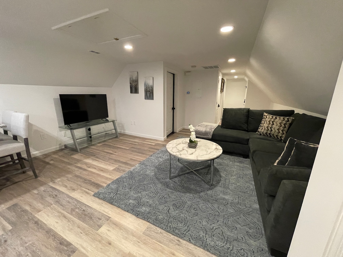 The living area features a comfortable sofa facing a 55-inch smart TV mounted on the wall. A round coffee table is positioned on a richly patterned area rug, with soft lighting illuminating the space. Nearby, a dining table with chairs is visible.