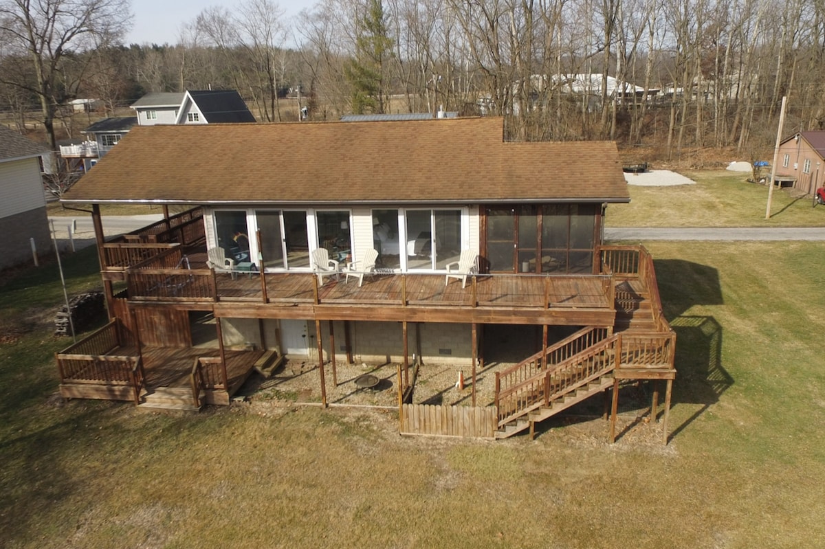 An aerial view of a spacious cottage features a large wooden deck wrapping around the building. The screened porch, visible at the front, is adorned with white chairs. Surrounding the cottage, the grassy yard is bordered by sparse trees, and nearby roads are also seen.