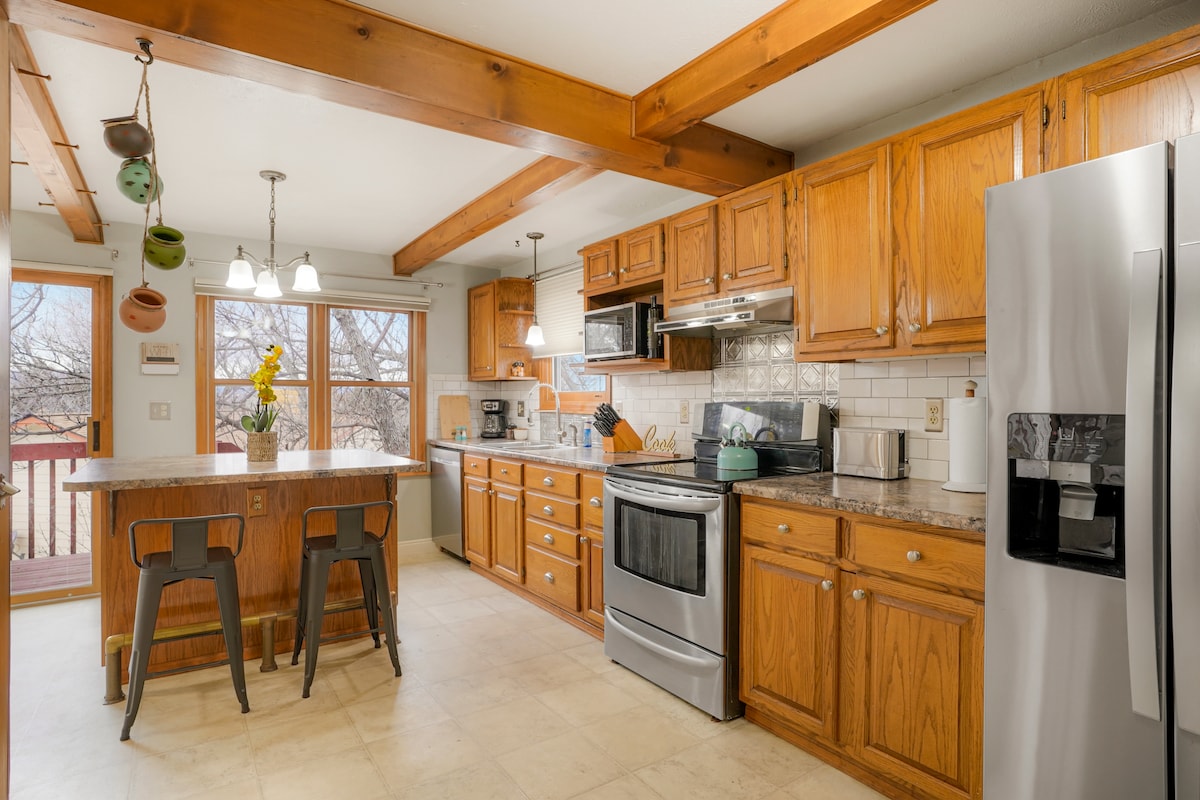 The kitchen features wooden cabinetry arranged along the walls with a countertop and stainless steel appliances. A central island with bar stools offers additional seating. Natural light enters through a window above the sink, with a nearby door leading to an outdoor deck.