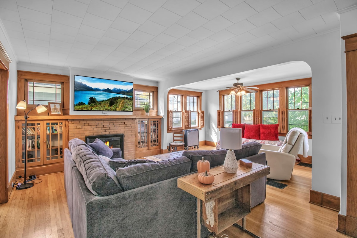 A spacious living area features comfortable seating arrangements, including a large sectional sofa. Natural light is invited in through multiple windows. A television is mounted above a brick fireplace, while light wood accents provide warmth to the room. Decorative plants and furniture enhance the inviting spaces.