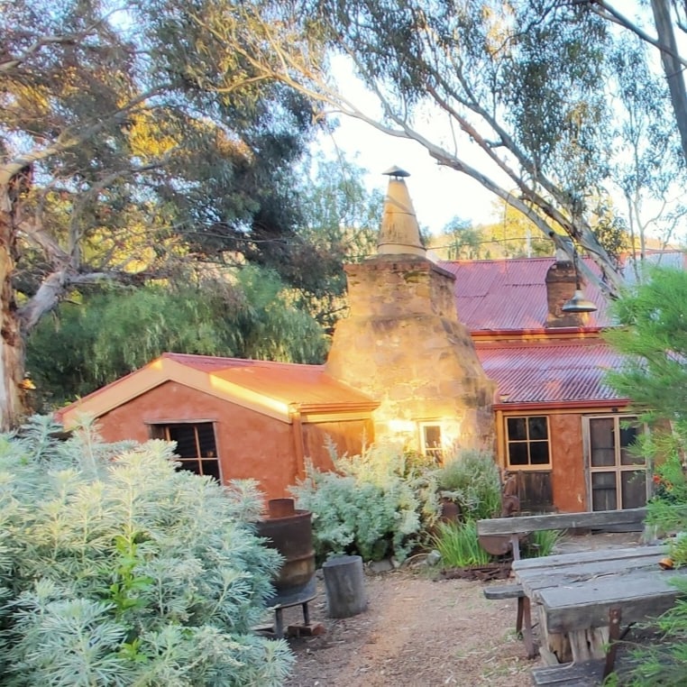 A rustic building with a warm earth-toned exterior is surrounded by lush greenery. Sunlight filters through the trees, illuminating the stone chimney and the red metal roof. Wooden tables are visible in the garden area, creating a serene outdoor space.