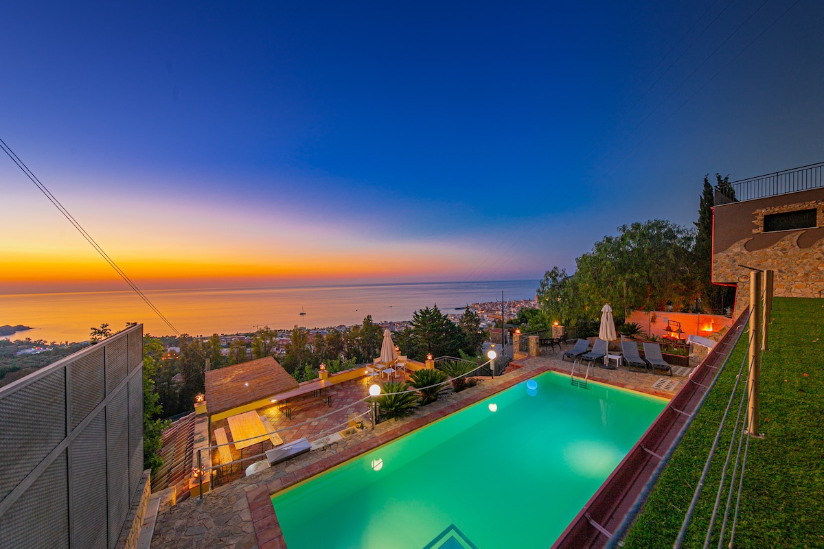A scenic view of a serene pool area is presented, with lounge chairs arranged around the illuminated turquoise water. The backdrop features a stunning sunset over the horizon, blending warm orange and blue tones above the coastal landscape.