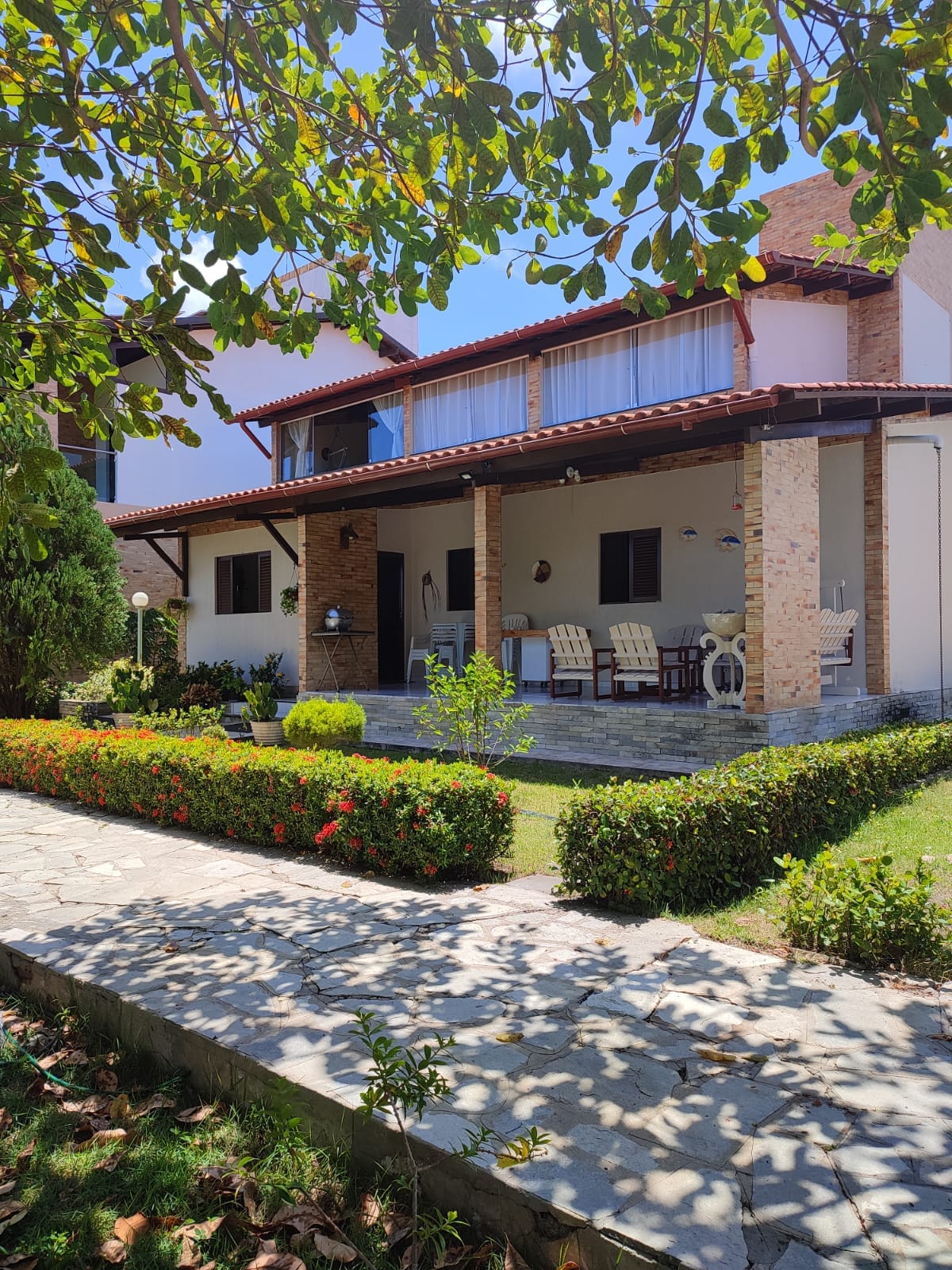 A serene exterior view of the accommodation is presented, featuring a porch shaded by trees and surrounded by manicured gardens. Comfortable seating is arranged under the overhang, with a stone pathway leading to the entrance. Bright sunlight filters through the foliage.