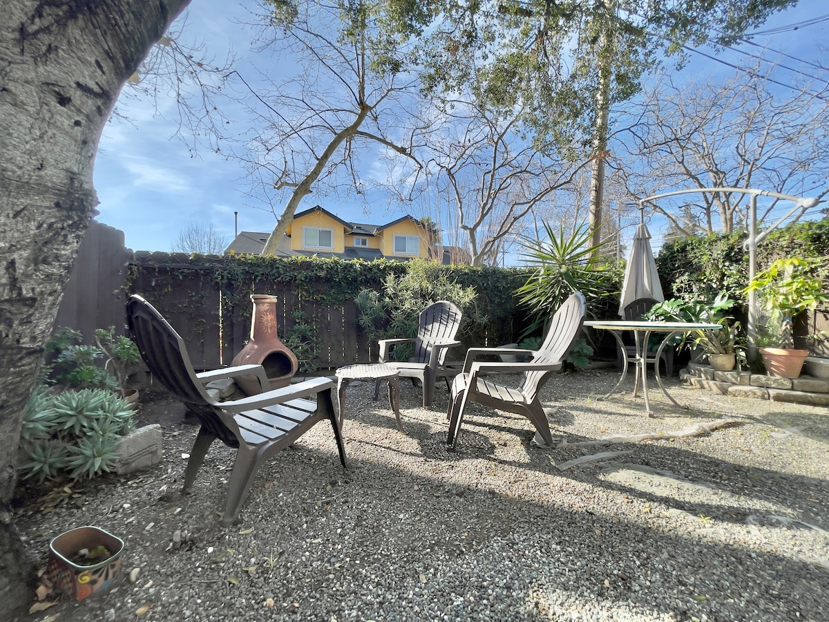 A garden area is highlighted with a gravel surface and several black outdoor chairs arranged around a small table. Nearby, a shaded area features a planted pot and a chiminea. Tall plants and trees create a natural border around the space.
