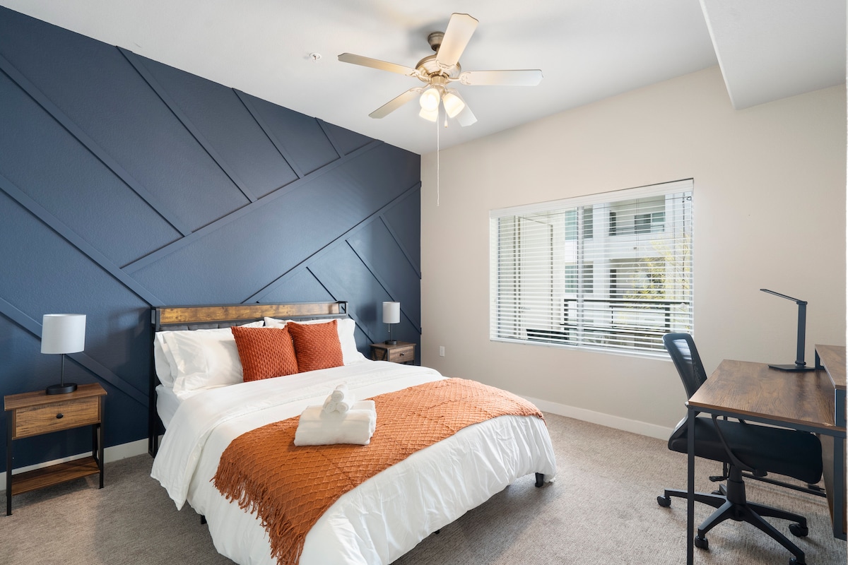 The main bedroom features an accent wall with a deep blue design. A queen bed is dressed in white linens, complemented by an orange throw. A window allows natural light and a desk with a chair is included for workspace convenience.