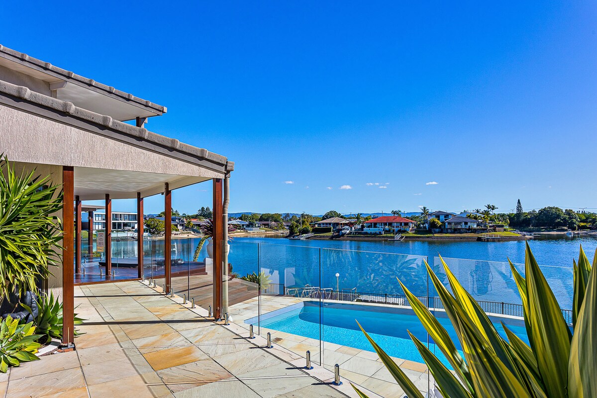 A serene waterfront view is captured from the spacious outdoor deck, featuring a large sun-drenched pool beside the water. Lush greenery frames the area, while the clear blue sky reflects over the tranquil waterway, offering a peaceful environment for relaxation.