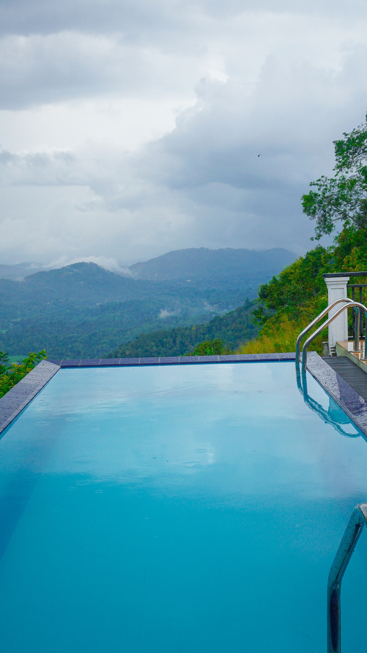 An infinity pool is seen, featuring clear blue water that merges with the horizon. Lush green mountains extend into the distance, partially obscured by soft clouds. The pool's edge is lined with stylish gray tiles and a metal railing on one side.