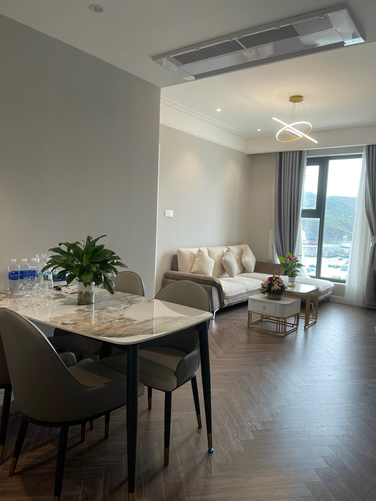 A modern living space features a dining table set for four, with a marble tabletop and elegant chairs. A cozy sofa with decorative pillows is positioned beside a large window, allowing natural light to illuminate the room. A potted plant and a small coffee table complete the setting.