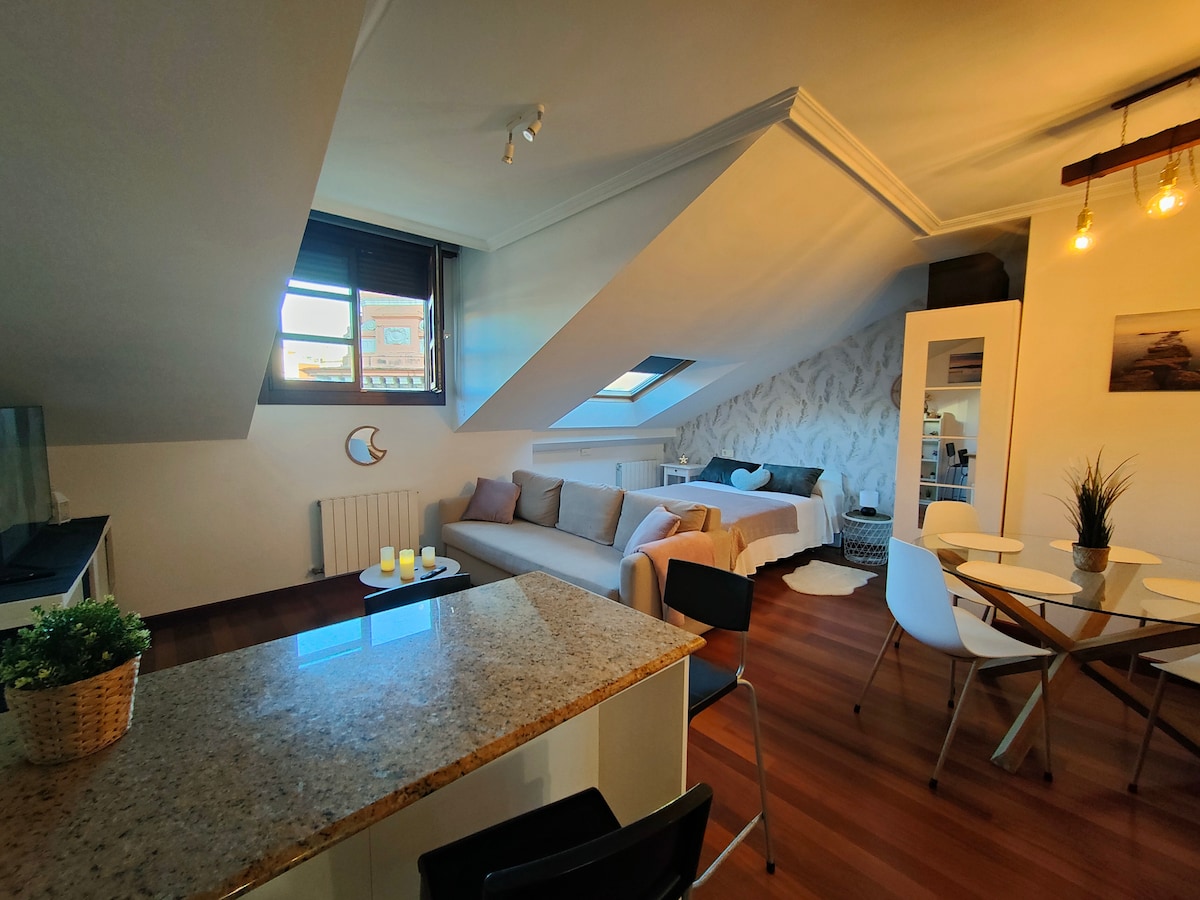 This inviting loft features a spacious living area with natural light streaming through the angled windows. A cozy seating arrangement is present alongside a dining area with white chairs. The kitchen island, topped with light-colored granite, offers a functional space for meal preparation.